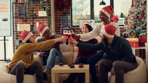ワインのグラスをクリックする人々の多民族グループは 冬の休日のイベントを祝うためにトーストを作ります オフィスでクリスマスの前夜祭の間にサンタの帽子と飲酒アルコールを身に着けている — ストック写真