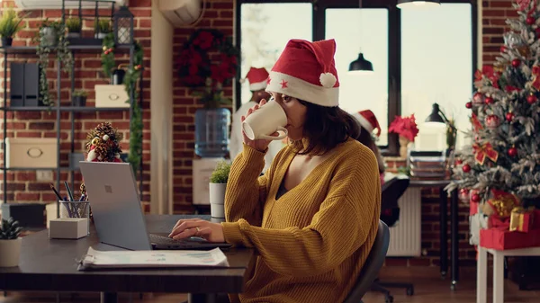 在圣诞节前夕 新员工在笔记本电脑上写报告 在办公室里工作 装饰着季节性的装饰品和灯光 圣诞佳节期间 头戴圣诞礼帽的节日女性发送电子邮件 — 图库照片