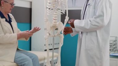 Sağlık kontrolünde insan iskeletini inceleyen doktor yaşlı hastaya anatomi kemik teşhisini açıklıyor. Ortopedik tedaviye yardımcı olmak için omurilik analizi yapıyorum..