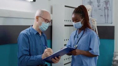 Hemşire, kabinede yaşlı bir hastayı muayene ediyor. Coronavirus salgını sırasında hastalara danışmanlık yapıyor. Tıbbi asistan ve yaşlı adam sağlık desteğinden bahsediyorlar..