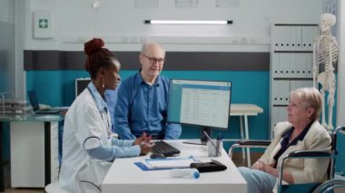 Female physician doing medical consultation with old woman in wheelchair, talking to patient and husband about disease treatment and recovery. Elderly person with chronic disability in office.