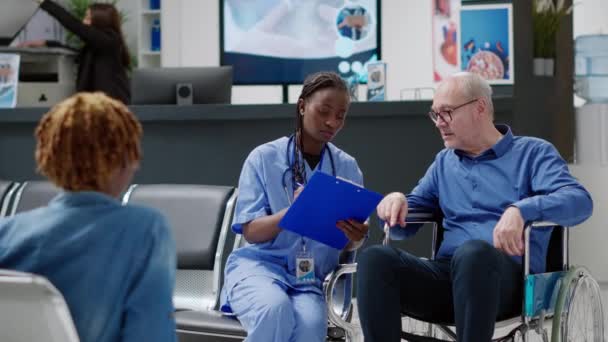 African American Nurse Consulting Senior Man Wheelchair Giving Treatment Medicine — Vídeo de stock