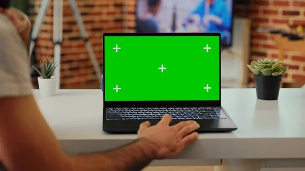 Male employee working with laptop and greenscreen template on display, looking at isolated mockup screen. Using blank chroma key copyspace background on wireless pc at home desk.