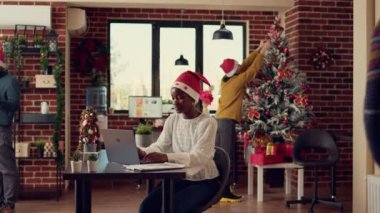 Afro-Amerikan bayram kadını dizüstü bilgisayarında veri tarıyor, Noel tatilinde ofiste Noel Baba şapkası takıyor. Çalışanlar kış geleneğini dekorasyon ve çalışmayla kutluyorlar..
