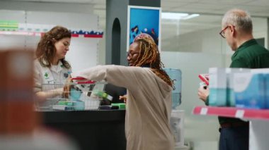 Eczacı, Afrikalı Amerikalı bir kadının eczaneden ilaç ve sağlık hizmeti almasına yardım ediyor. Çalışanlar ilaç satmak için ilaç ve vitamin kutularını tarıyor..