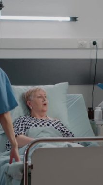 Dikey video: Hastane koğuşunda emekli bir hastayla istişarede bulunan doktor. Doktor hasta kadınla konuşurken hemşire de tıbbi muayene ve iyileşme için destek veriyor. Tıbbi