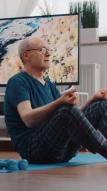 Dikey video: Nilüfer pozisyonunda oturan yaşlı adam yoga minderinde meditasyon yapıyor. Zen emeklisi egzersiz yaptıktan sonra rahatlamak ve meditasyon yapmak için nefes alıyor. Sağlıklı olmak için meditasyon yapan yaşlı bir insan.