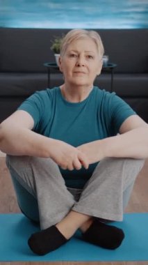 Dikey video: Evde egzersiz yapmaya hazırlanan yaşlı kadının portresi. Egzersiz yapmak ve eğitim ekipmanlarıyla spor yapmak için yoga minderinde oturan yaşlı bir insan. Sportif aktif emekli