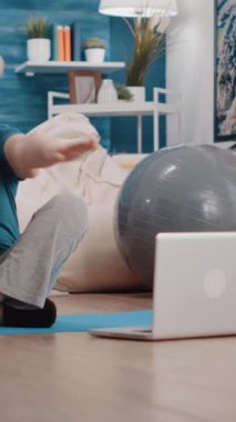 Dikey video: Evde yoga minderinde egzersiz yapan yaşlı kadın, dizüstü bilgisayardaki çevrimiçi eğitim dersinden sonra. Zeminde fiziksel egzersiz yapmak için bilgisayarda spor videosu izleyen yaşlılar..