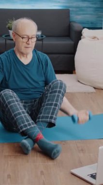 Dikey video: Üst düzey yetişkin dizüstü bilgisayarda egzersiz videosu izlerken fiziksel egzersiz yapmak için dambıl kullanıyor. Çevrimiçi eğitim dersini takip eden yaşlı bir adam bilgisayar ve ağırlık kaldırarak spor yapıyor.