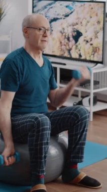 Dikey video: Son sınıf öğrencisi halterleri kaldırıyor ve beden eğitimi ve egzersiz yapmak için spor topunun üzerine oturuyor. Emekli insan evde kol kaslarını çalıştırmak için ağırlıklarla egzersiz yapıyor..