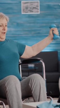 Dikey video: Yaşlı yetişkin tekerlekli sandalyede oturuyor ve tablet üzerinde eğitim videosunu takip ederken ağırlıklarla egzersiz yapıyor. Engelli yaşlı kadın, internet dersinde kullanacağı dambılları kullanıyor..