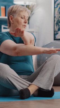 Dikey video: Yaşlanan kişi fiziksel egzersiz yapıyor ve tablet üzerinde egzersiz videosu izliyor. Kıdemli bir kadın spor için online eğitim dersini takip ediyor, yoga minderinde oturuyor. Emekliler evde egzersiz yapıyor.