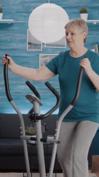 垂直ビデオ 自宅でカーディオマシンで物理的な運動を行う年金受給者 固定式電動自転車を使用してトレーニングを行うシニア女性 設備でフィットネス活動を行う者 — ストック動画