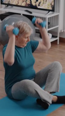 Dikey video: Üst düzey kadın ağırlık kaldırmak için dizüstü bilgisayar kullanıyor, online egzersiz dersi videosu izliyor. Emeklinin koçla antrenman yaparken fiziksel egzersiz yapması.