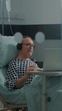 Dikey video: Hastane koğuşundaki hasta klinikteki yoğun bakım odasında müzik dinliyor. Yaşlı adam hastalıkları ve hastalıkları tedavi etmek için tedavi görürken dinlenmek için dizüstü bilgisayar ve kulaklık kullanıyor.