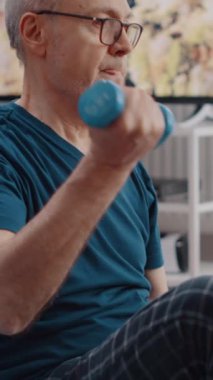 Dikey video: Halterli yaşlı insan eğitimini kapatın, yoga minderinde oturun. Emekli adam ağırlık kaldırıyor ve kol kaslarını çalıştırmak için fiziksel egzersiz yapıyor. Yetişkin esneme hareketleri ve...