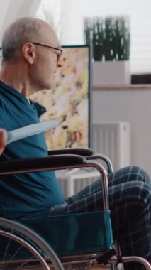 Dikey video: Emekli adam tekerlekli sandalyede oturuyor ve tablet üzerinde egzersiz videosu izlerken egzersiz yapmak için direniş bandını kullanıyor. Fiziksel engelli yaşlı yetişkin esnemek için elastik kemer çekiyor