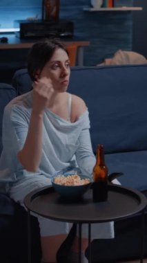 Dikey video: Şaşırmış bir kadın TV 'de film izlerken yüz ifadesine şaşırmış, koltukta patlamış mısır yiyor. Gece geç saatlere kadar televizyon seyreden yalnız bir genç bayan.