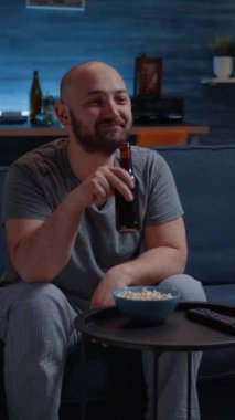 Dikey video: Komik adam gülerek TV 'de komedi filmi izliyor patlamış mısır yiyor ve bira içiyor. Evde yalnız başına heyecanlı, pijama giymiş bir şekilde rahat bir kanepeye oturmuş akşamın tadını çıkarıyor.