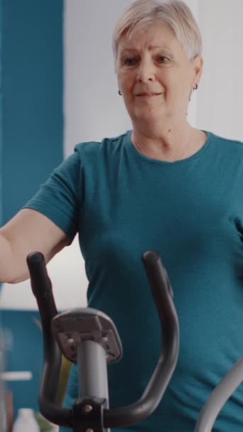 垂直ビデオ 自宅で静止自転車に乗って引退した女性の閉じる カーディオマシンを使用している高齢者は 静的自転車でサイクルし 物理的な運動を行う 年金生活とフィットネスを行う — ストック動画