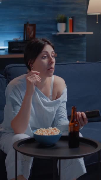 垂直ビデオ 退屈女性は夜遅くにポップコーンを食べるリモートコントローラを保持し テレビチャンネルを切り替えるエンターテイメントテレビを検索します 女性リラックスした座席でソファでPajama飲料ビール — ストック動画