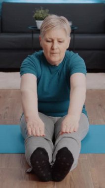 Dikey video: Yaşlı kadın yoga minderinde aerobik pozisyonu alıyor. Fiziksel egzersiz yapan ve sağlık ve sağlık eğitimi alan yaşlı bir insan. Emekliler evde jimnastik yapıyor..