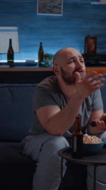 Dikey video: Kızgın hayal kırıklığına uğramış spor hayranı favori takımın kaybetmesini izliyor, fotbol yarışması sırasında televizyonda bağırıyor. Evde yalnız başına üzgün bir adam geceyi kanepede oturmuş oyun takımını destekleyerek geçiriyor.