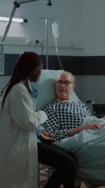 Dikey video: Afro-amerikan doktor hasta hasta hasta ile hastanede konuşuyor sağlık sorunları ve yaralanmaları için. Yaşlı adam doktor ve hemşireye yoğun bakım ve hastalıkları soruyor.