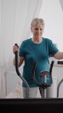 Dikey video: Yaşlı bir kadın evde egzersiz yapmak için sabit bisiklet kullanıyor. Emekli bir yetişkin, elektrikli bisiklet ve kardiyo makinesiyle fiziksel egzersiz yapıyor. Spor aletleri ile son sınıf öğrencisi..