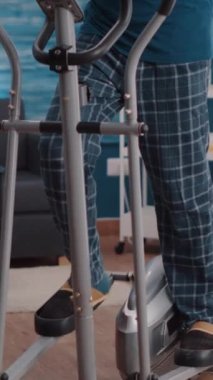 Dikey video: Dayanıklılık ve spor için yaşlı bir adam sabit bisiklete biniyor. Son sınıf öğrencisi statik bisikleti evde egzersiz yapmak ve kas çalıştırmak için kullanıyor. Emekli yetişkin bisikleti.