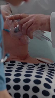 Dikey video: Doktorun hastaya oksijen maskesi takarken hemşirenin sağlık hizmetlerine destek vermesi. Hastane koğuşunda tıbbi solunum sorunları için yardım alan emekli kadın