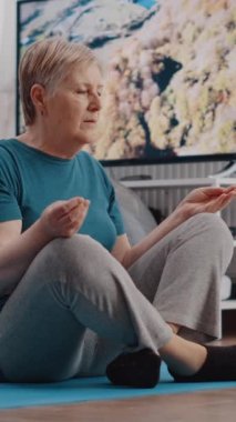 Dikey video: Yaşlı kişi evde meditasyon yapmak için yoga minderinin üzerinde lotus pozisyonunda oturuyor. Son sınıf öğrencisi kadın sakin ve dengeli olmak için zen meditasyonu yapıyor fitness eğitiminden sonra dinleniyor. Emekliler meditasyon yapıyor.