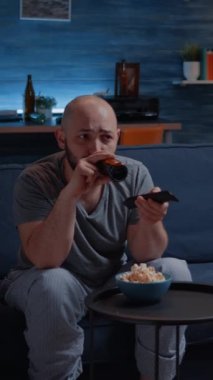 Dikey video: Sıkıcı adam televizyon kanallarını değiştiriyor rahat koltukta oturup bira içiyor, gece geç saatlerde evde tek başına oturuyor. Televizyon seyreden biri elinde uzaktan kumandayla bir komedi arıyor.