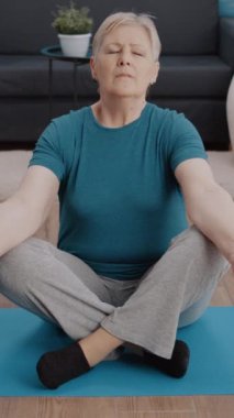 Dikey video: Yoga minderinde meditasyon yapmak için Lotus pozisyonunda oturan gözleri kapalı emekli kadın. Yaşlı insan denge ve ahengi bulmak için meditasyon yapıyor, aktivitelerle rahatlıyor. Fitness eğitimi