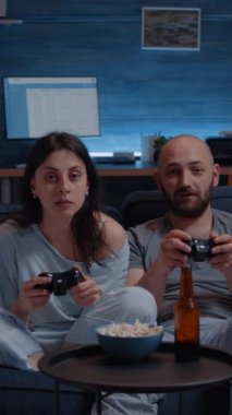 Dikey video: Odaklanmış kararlı çift gece geç saatlerde bilgisayar oyunu oynayarak online yarışmayı kazanmaya çalışıyor. Kanepede oturup, joystick kullanarak video oyunu oynayan oyuncular.