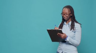 Afro-Amerikalı kadın kağıtlara yazı yazıyor. Dosyaları mavi arkaplanda not almak için panoya kullanıyor. Belgeler ve evraklar hakkında bilgi veren, neşeli hisseden biri..