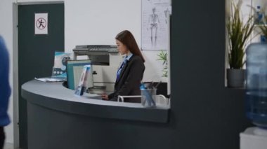 Asyalı resepsiyonist, sağlık sigortası için hastane kayıt masasında bilgisayar kullanıyor. Sağlık personeli lobideki resepsiyon masasında check-up randevuları üzerinde çalışıyor. El kamerasıyla..