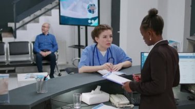 Hemşire ve resepsiyonist, resepsiyondaki check-up kağıtlarını inceliyor, hastalara yardım etmek için sağlık sigortası üzerinde çalışıyor. Sağlık personeli rapor formları ve istişareler hakkında konuşuyor..