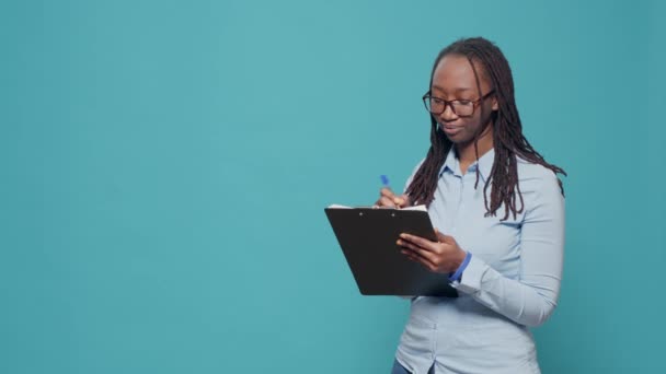 非裔美国妇女在纸上书写文字 用剪贴板上的文件记录蓝色背景 在文件和文书工作中提供信息的人 感到快乐 — 图库视频影像