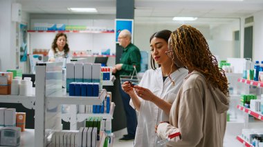 Çok ırklı bir grup müşteriye eczaneden ilaç ve vitamin almalarında yardım eden bir tıp çalışanı raflardaki haplardan bahsediyor. İlaç ürünleri ve ilaçlara yardım etme.