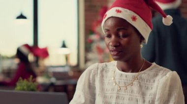 Şenlik ofisindeki dizüstü bilgisayarında Noel ağacı ve ışıklarıyla Noel Baba şapkası takan bir kadın işçi. Mevsimsel kış tatili boyunca rapor üzerinde çalışan bir kadın. Süslü bir yer. Üçayak atışı.