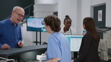 Hastane resepsiyonunda çalışan çeşitli tıbbi ekipler, sağlık kontrolleri için rapor veren yaşlı bir hastaya yardım eden hemşire. Klinik sağlık personeli kayıt masasında. El kamerasıyla..