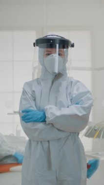 Dikey video: Ağız sağlığı kliniğinde virüs koruması için koruyucu elbisenin içinde duran dişçi portresi. Beyaz elbiseli kadın dişçilik ameliyatı ve cerrahi konsültasyon için hazırlandı.