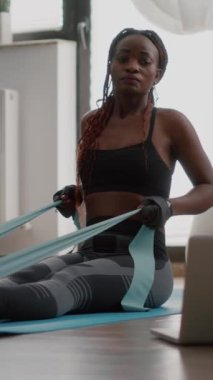 Verical video: Spor giyim bölümünde formda siyah bir kadın dizüstü bilgisayarda vücut kaslarını çalıştırmak için spor yaparken sağlıklı yaşam tarzının keyfini çıkarıyor. Yetişkin sporcu oturma odasında yoga haritasında oturuyor.