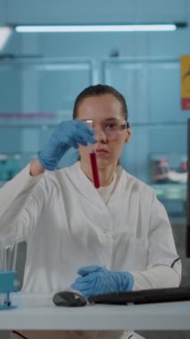 Dikey video: Laboratuvarda bilim deneyi için test tüpündeki maddeleri analiz eden kadın kimyager. Koruyucu gözlüklü ve eldivenli biyoloji uzmanı yenilikler için şişe ve laboratuvar malzemesi kullanıyor..