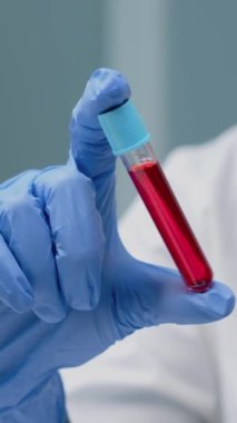 Dikey video: Laboratuvarda incelenmek üzere kanla doldurulmuş boşta gezenin kapanışı. Doktor tarafından bilim için kullanılan sıvı çözeltisi ile şeffaf cam araçlara bakan eldivenli bilim adamı