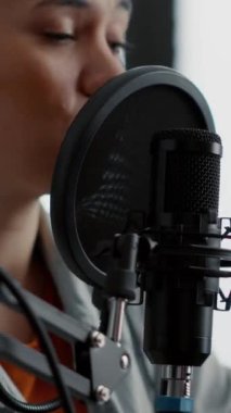 Dikey video: Genç bir kadın profesyonel mikrofon kullanırken izleyicilerle konuşuyor. İnternet canlı radyo programı sunucusu modern kulaklıklarla stüdyoda otururken günün son haberlerini sunuyor..