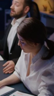 Dikey video: Kameraya bakan gülümseyen iş kadınının portresi iş dünyasının ofis odasında gece geç saatlerde fazla mesai yapıyor. İşkolikler çeşitli çok ırklı takım çalışmalarını tartışıyor