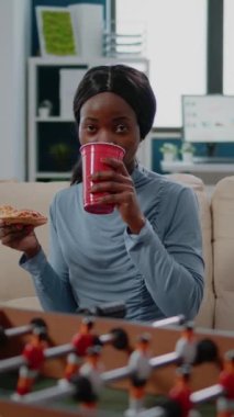 Dikey video: İşten sonra eğlenmek için elinde bir dilim pizza ve birayla gezen kadının portresi. Afrika kökenli Amerikalı yetişkinler iş arkadaşlarıyla birlikte langırt masasında eğleniyorlar.